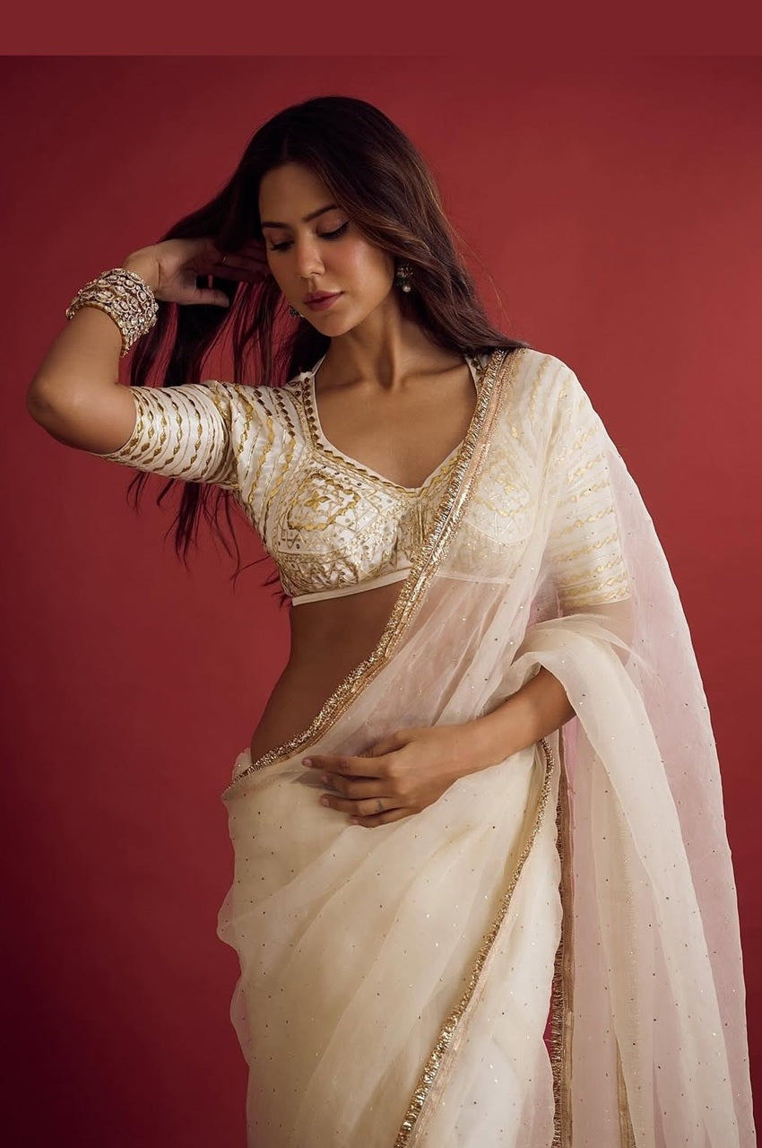 Woman wearing a traditional cream saree with gold embroidery against a red background