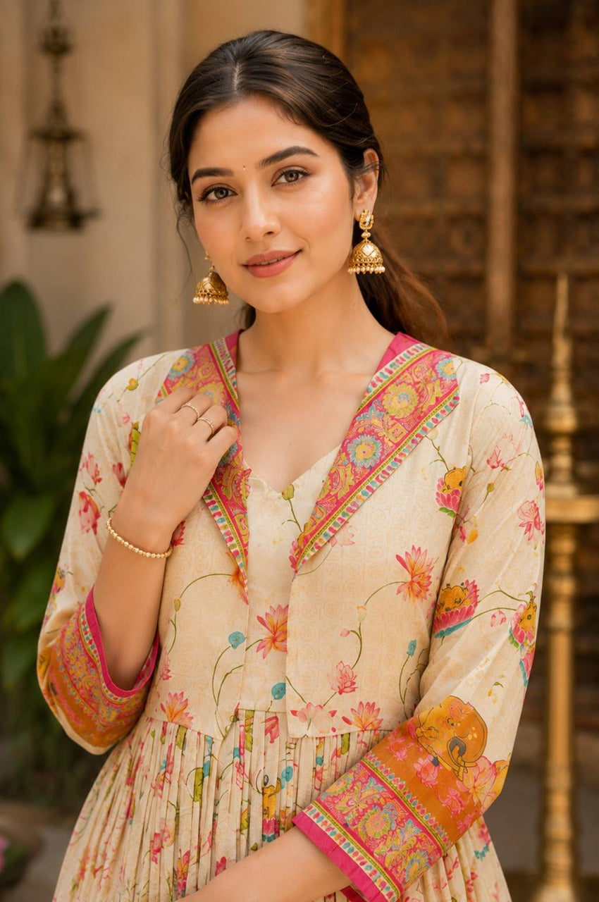A woman wearing a beige printed kurta with colorful floral motifs and ornate pink and orange border details, styled with gold earrings and bracelets, set against a warm, blurred background.