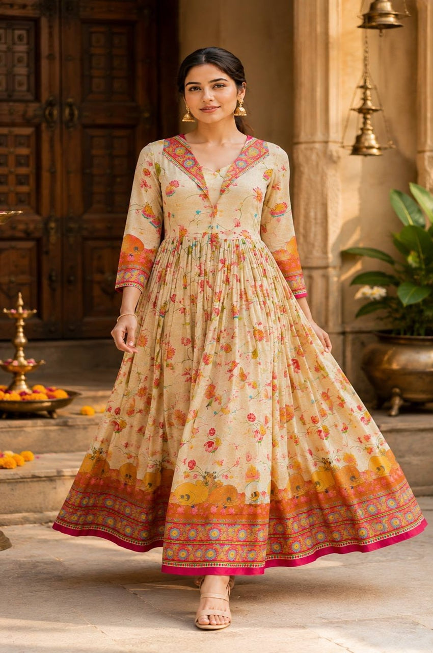 A woman models a beige printed maxi dress with orange, pink, and yellow floral motifs and a patterned border, standing in front of a traditional stone building with wooden doors.