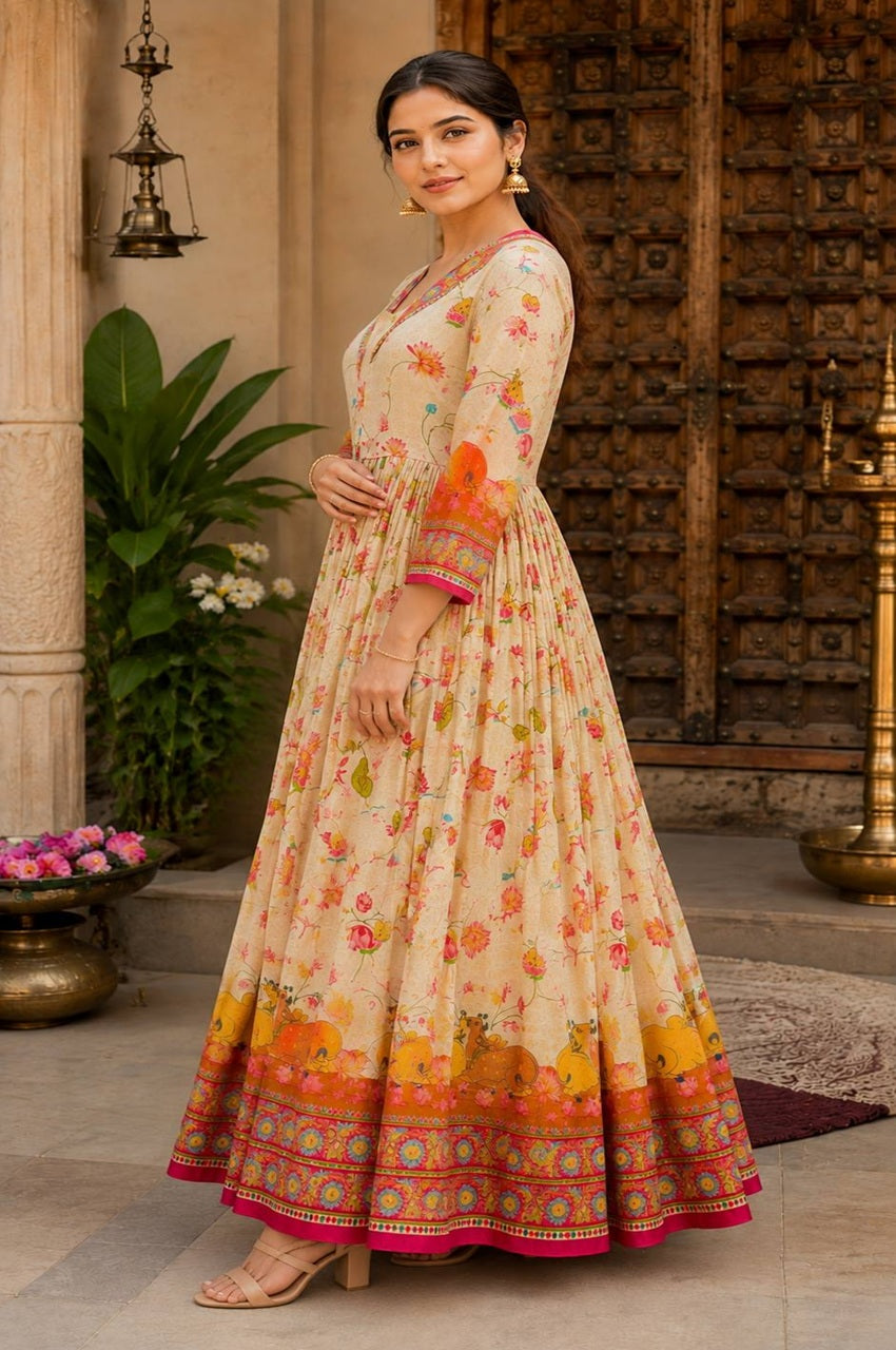 A woman models a long cream-colored printed Anarkali gown with orange, pink, and yellow floral motifs and a decorative border, set against a traditional courtyard background.
