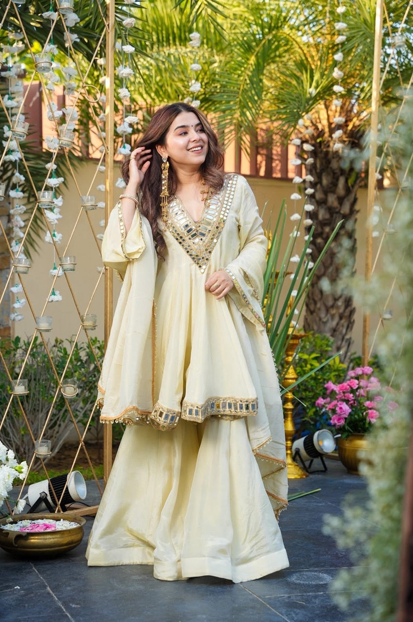 Woman in a cream traditional outfit standing in an outdoor setting with plants and decorations.