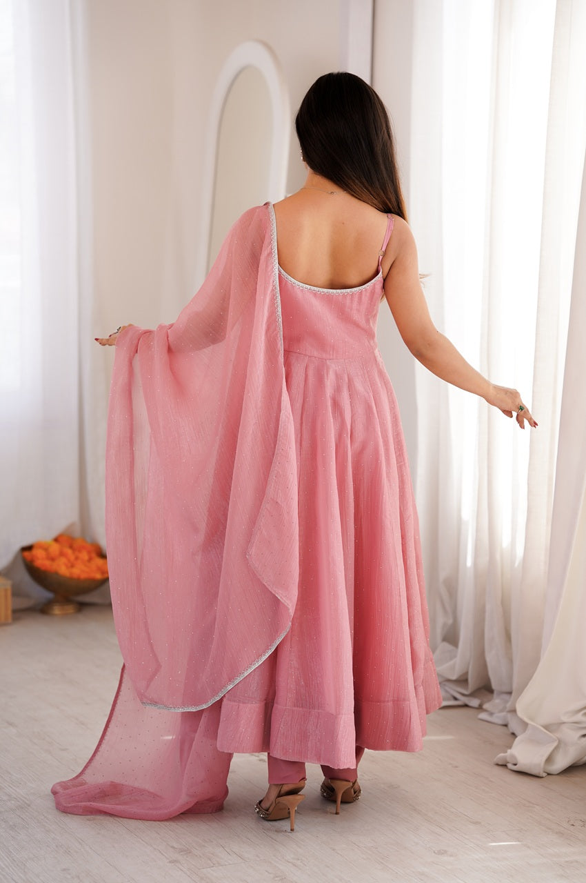 A woman viewed from behind wearing a pink sleeveless dress with a matching sheer pink dupatta, standing in a bright white room with curtains and a mirror in the background.