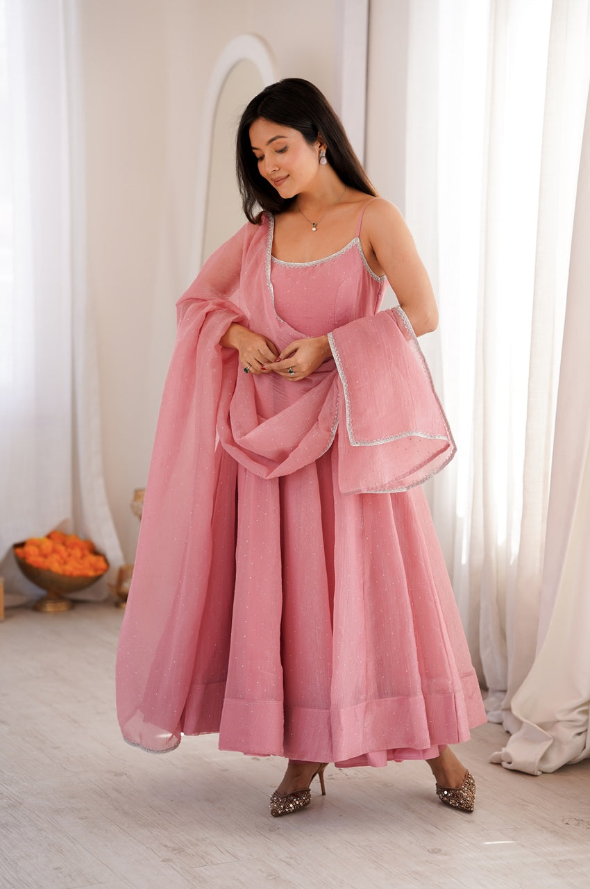 A woman modeling a dusty pink traditional outfit consisting of a sleeveless dress, a matching dupatta, and a long pink shawl with white border trim, set against a bright, neutral indoor background.
