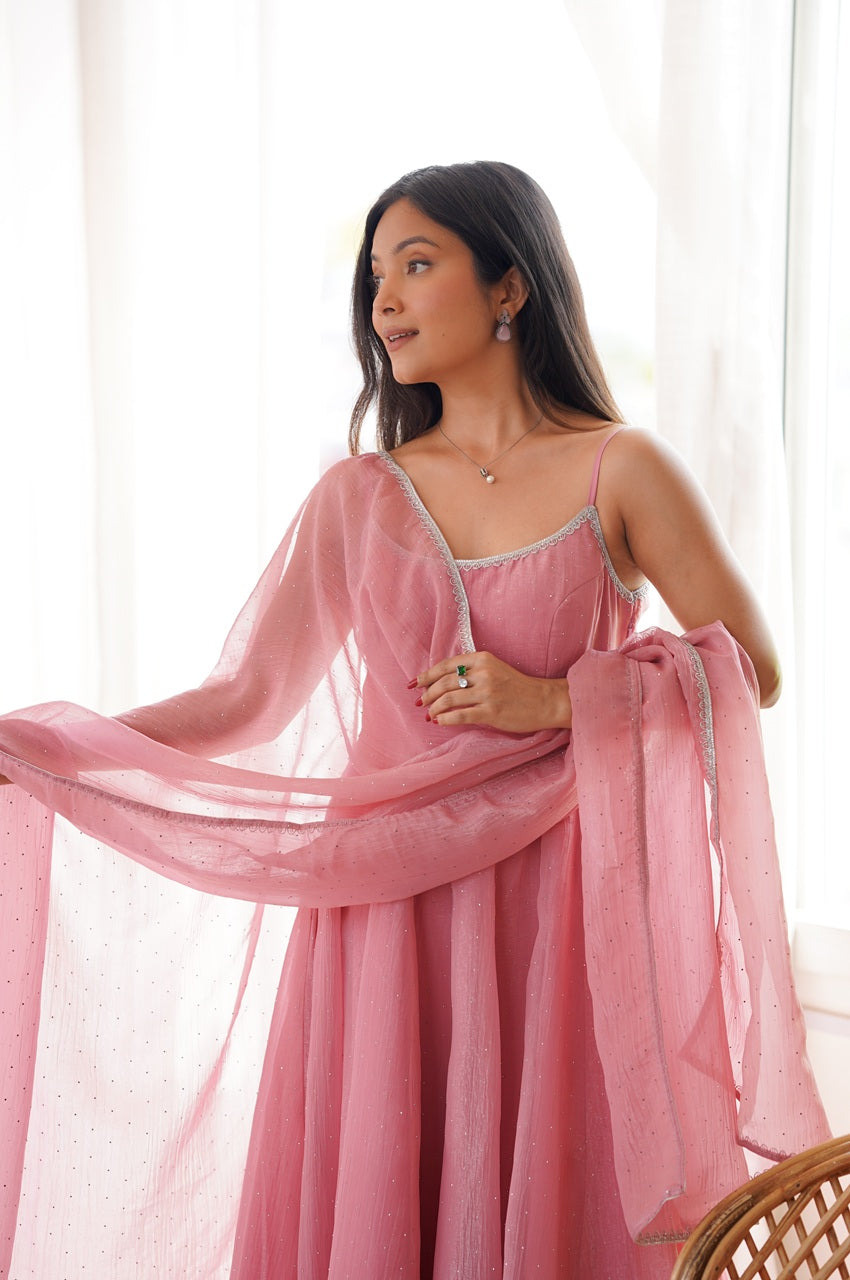A woman wearing a flowing dusty pink traditional dress with a matching sheer dupatta, standing in front of a bright window with white curtains.