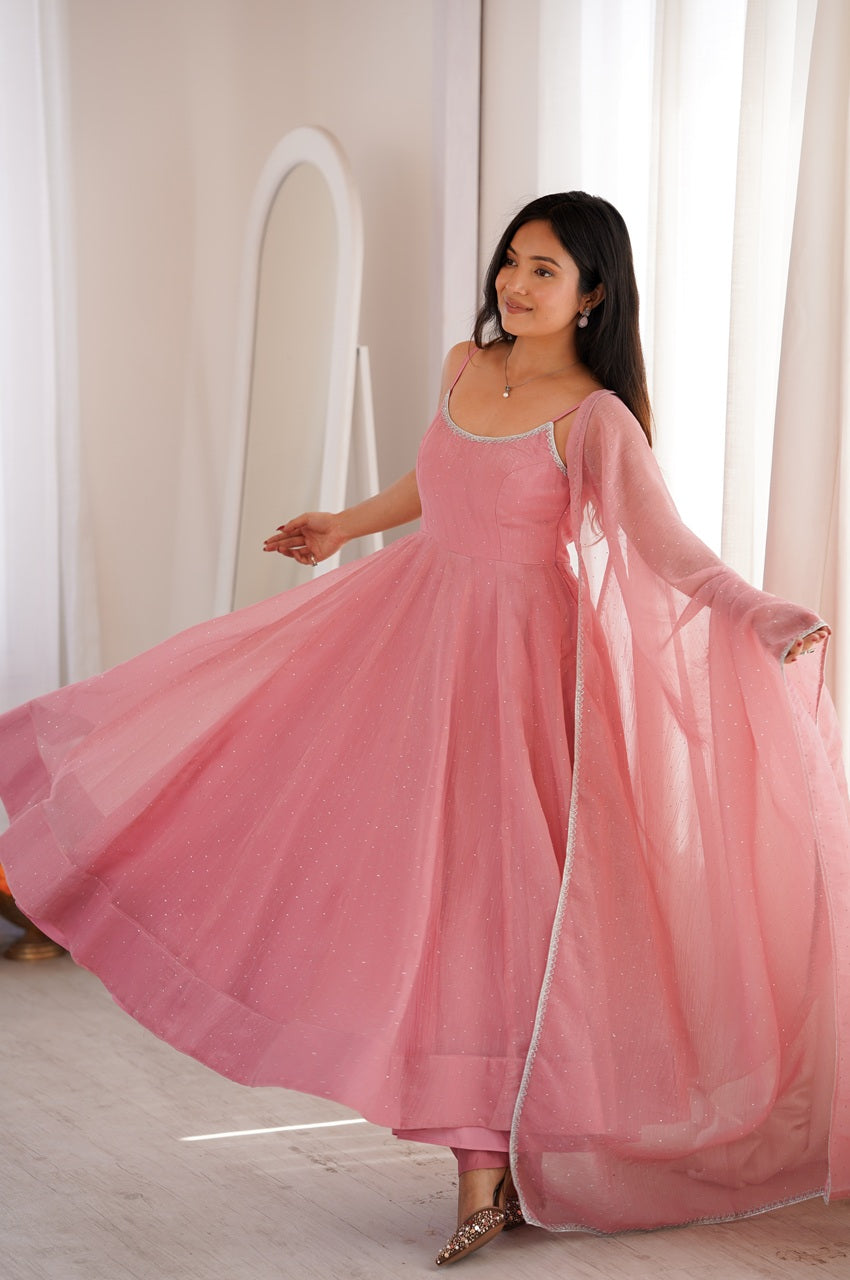 A woman wearing a flowing pink traditional dress with a sheer long overlay, captured mid-twirl in a bright indoor setting with white curtains and a mirror.