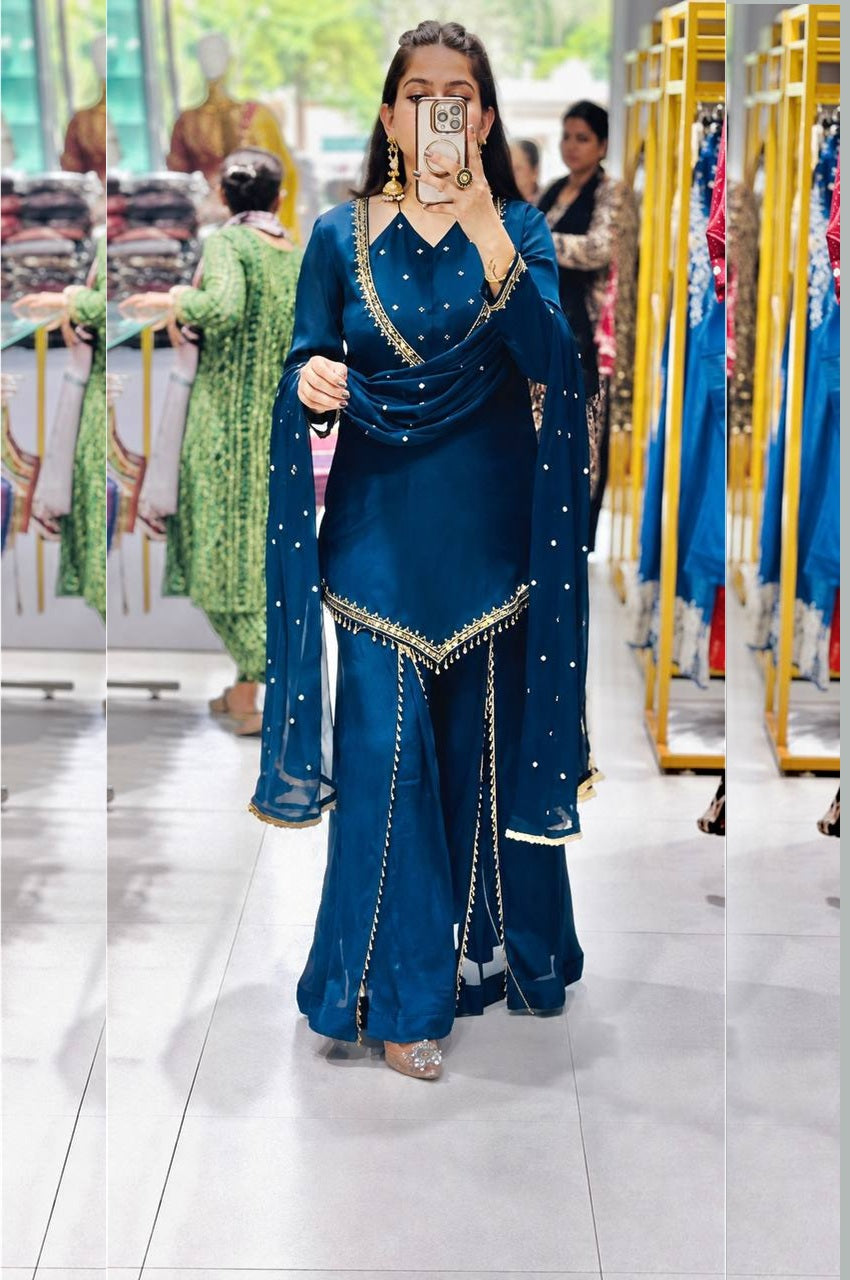 A woman takes a mirror selfie in a clothing store wearing a deep teal blue traditional outfit with gold embroidery and a matching dupatta.