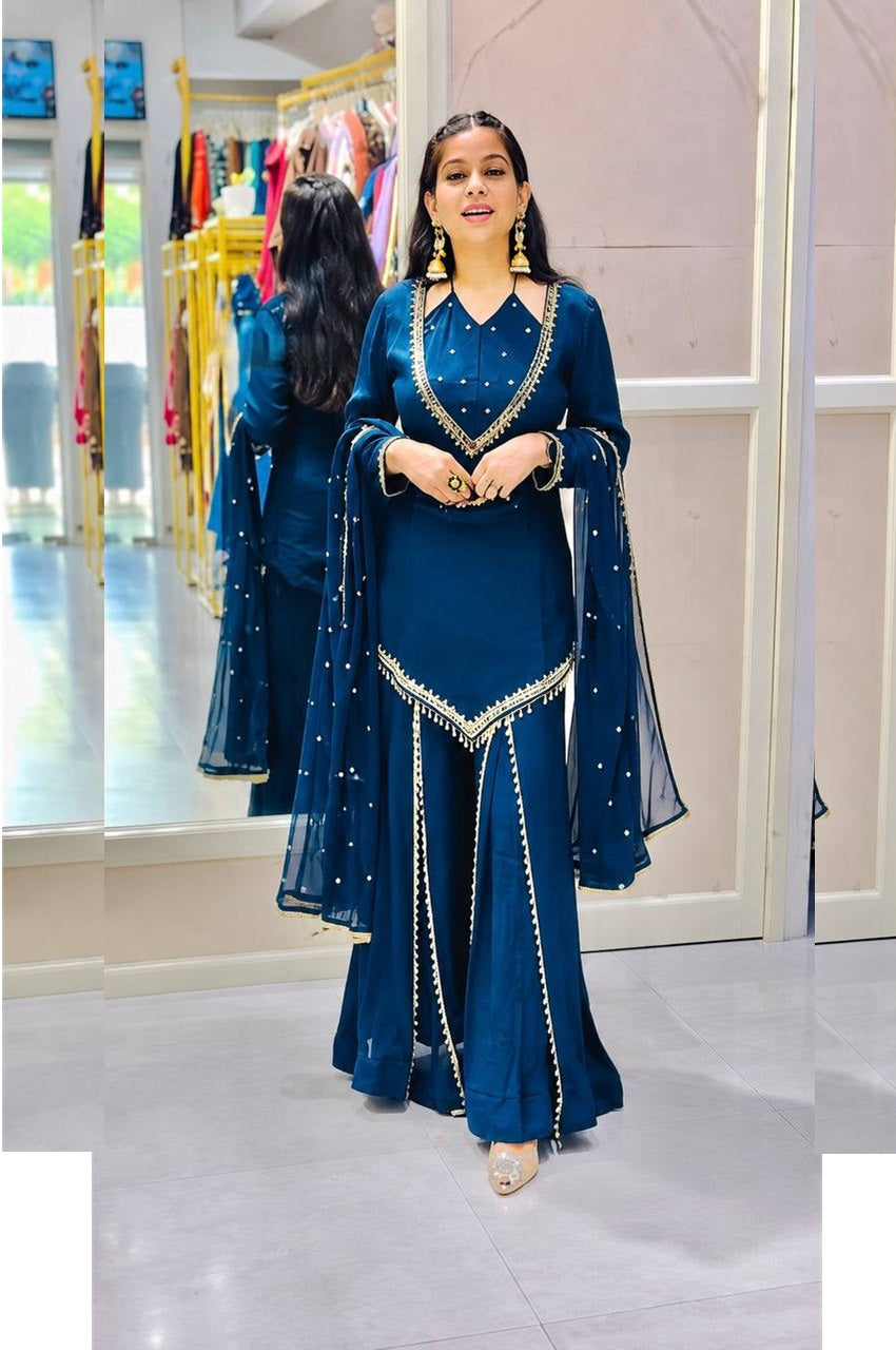 A woman modeling a deep blue traditional outfit with gold embroidery and a matching sheer dupatta in a boutique setting.