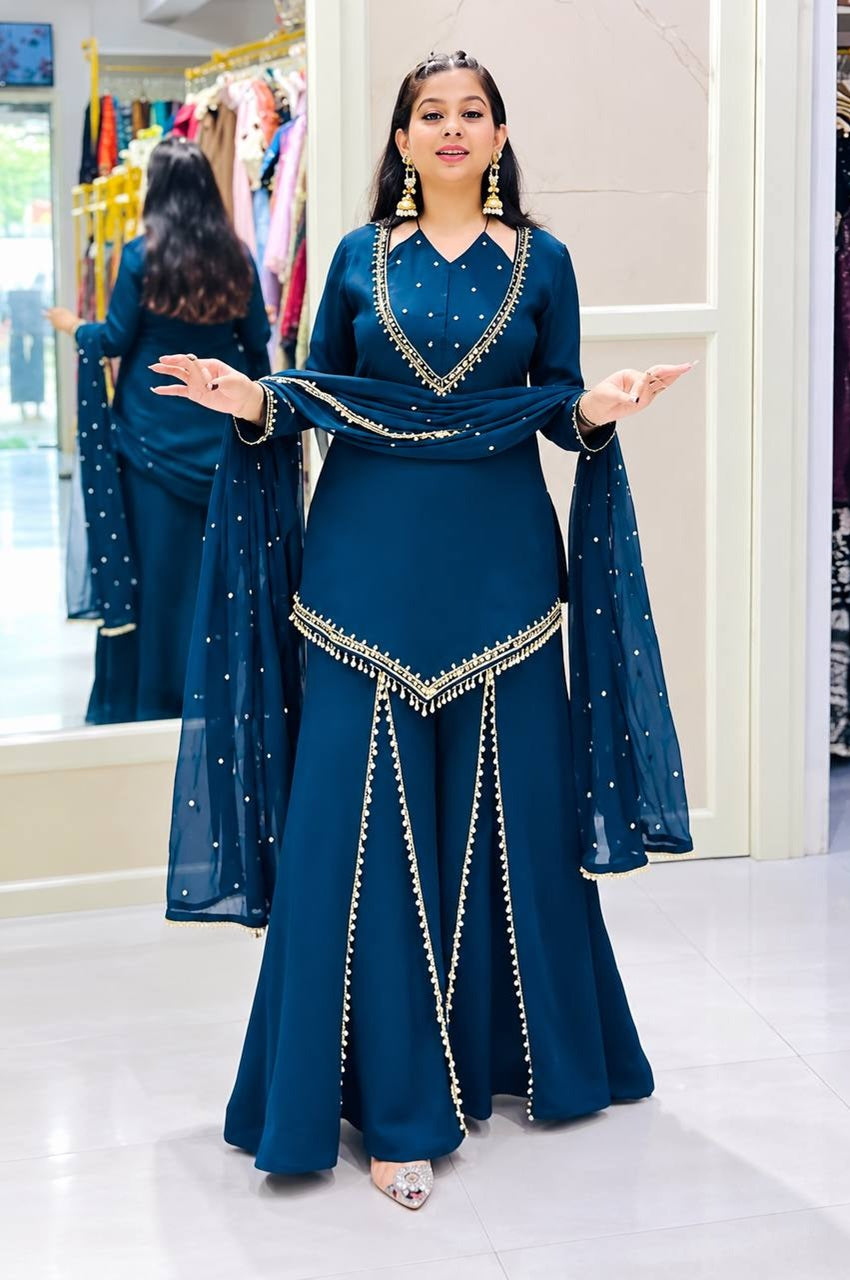 A woman modeling a deep teal blue traditional Indian outfit with gold embroidery, standing in a boutique. The outfit features a long-sleeved kurta with a V-neckline, flared pants, and a matching dupatta draped over her arms.