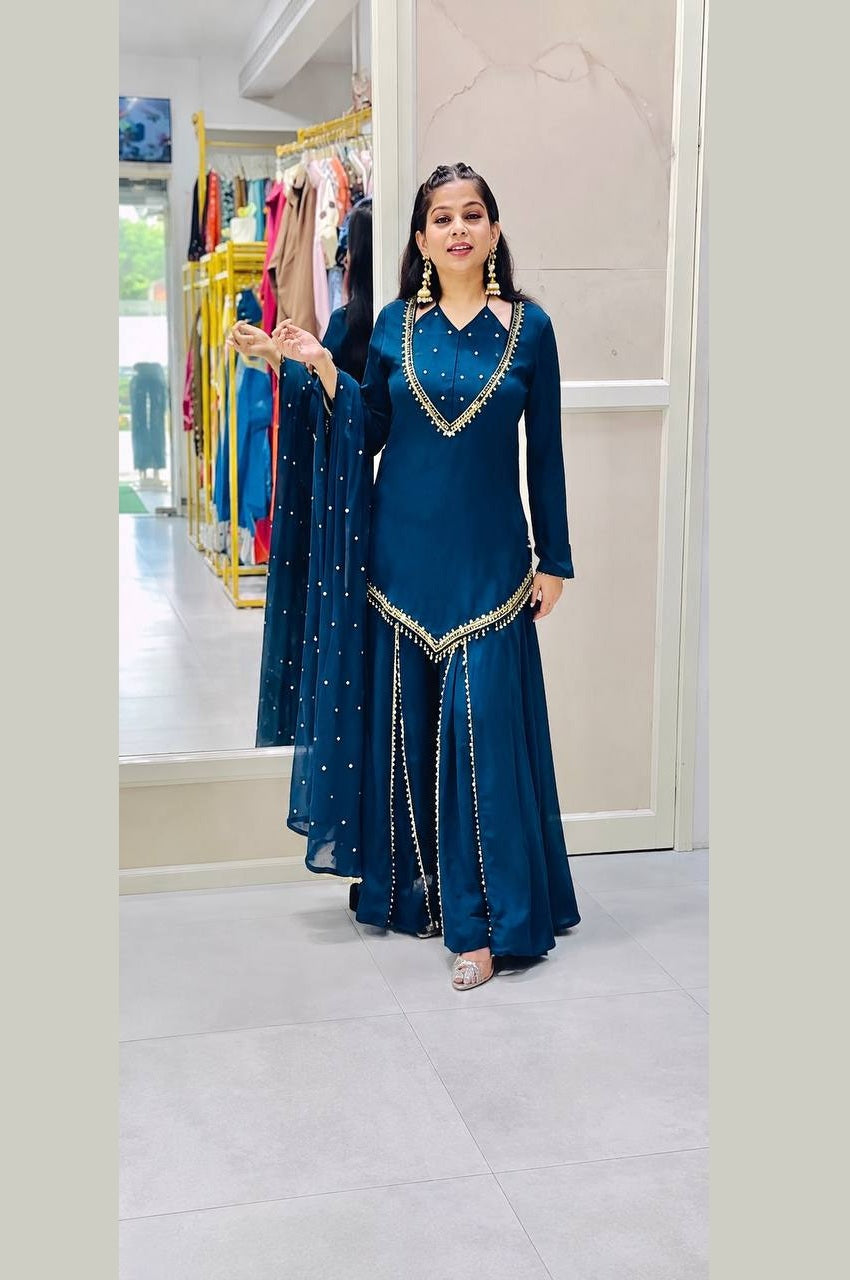 A woman modeling a deep teal blue traditional Indian outfit with gold embroidery, standing in a clothing boutique.