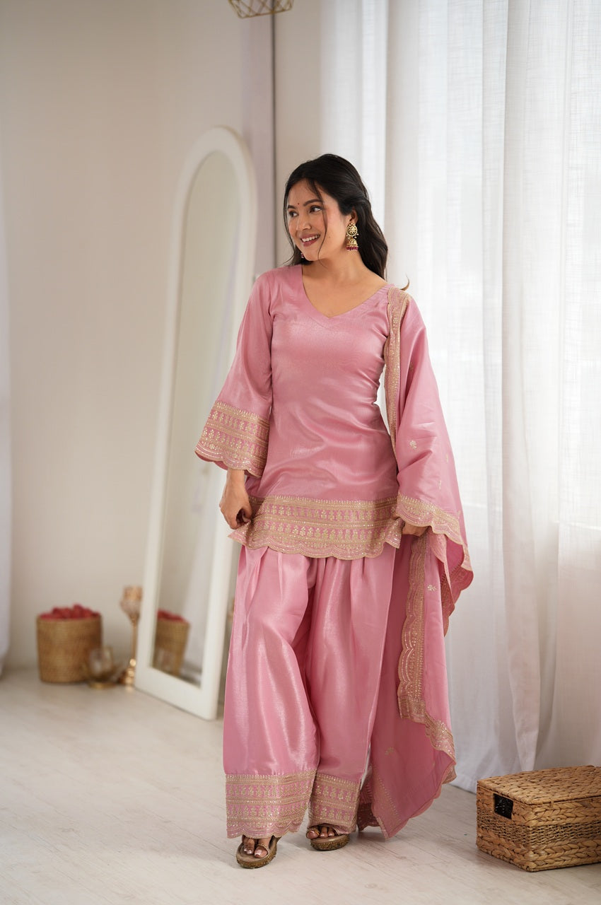 A woman modeling a pink traditional three-piece outfit with beige embroidered details, standing in a bright, minimalist room with white curtains and a floor mirror.