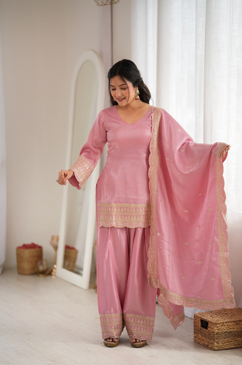 A woman modeling a pink three-piece traditional outfit with matching dupatta in a bright, neutral indoor room. The set includes a long-sleeve kurta with gold embroidery, matching wide-leg pants, and a sheer pink dupatta with a gold border.