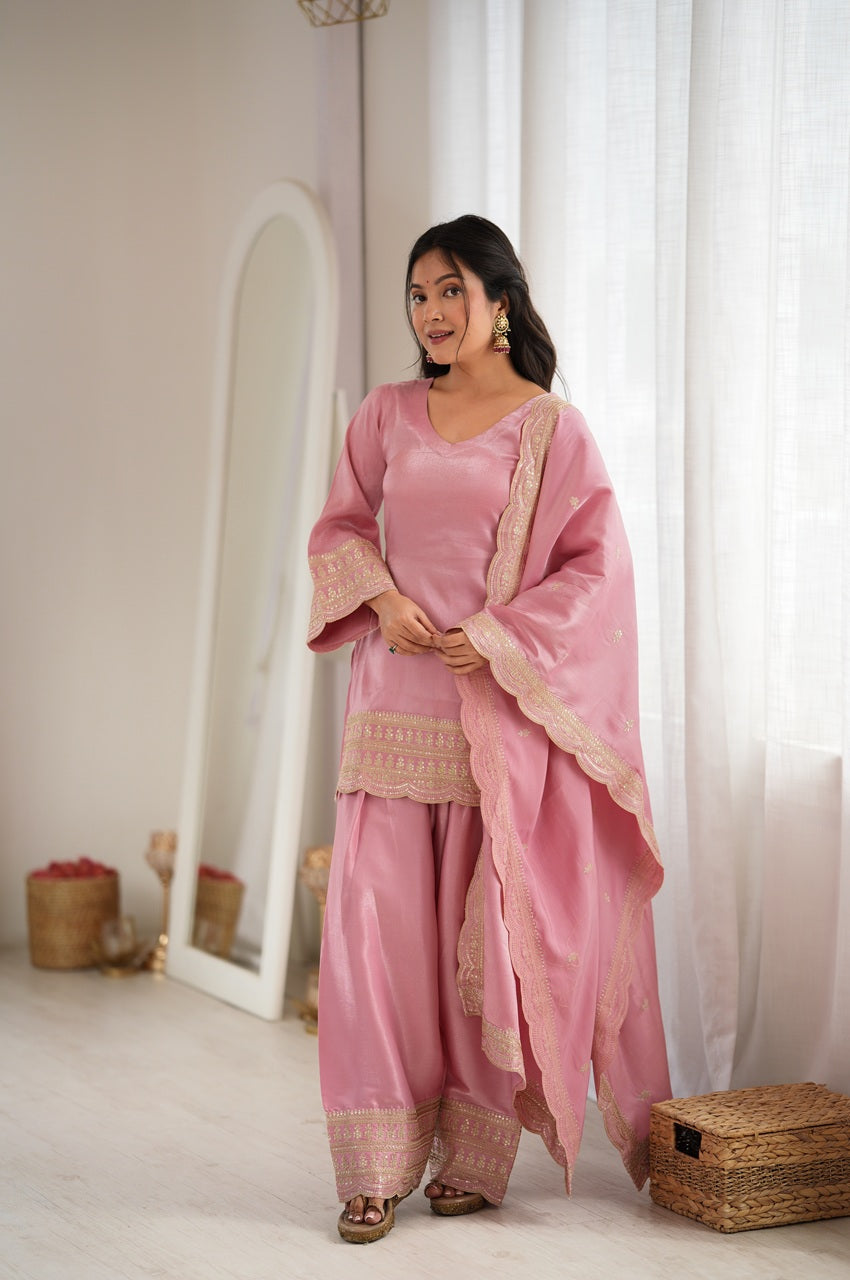 A woman modeling a pink traditional three-piece outfit with beige embroidered borders, standing in a bright, minimalist room with white curtains and a floor mirror.