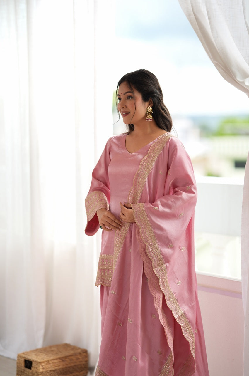 A woman wearing a pink traditional outfit with gold embroidered borders stands in front of a bright window with white curtains.