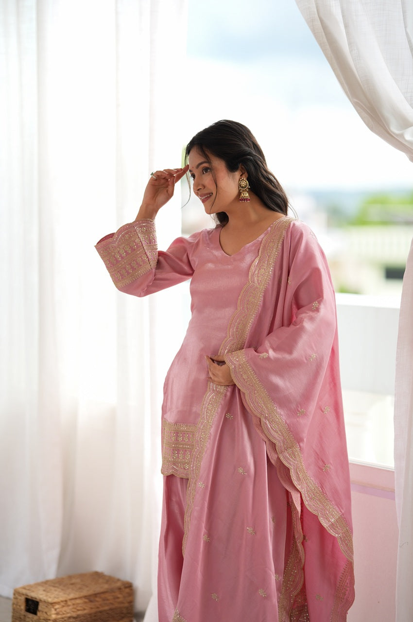 A woman wearing a light pink traditional outfit with gold embroidered borders, standing in front of a bright window with white sheer curtains.