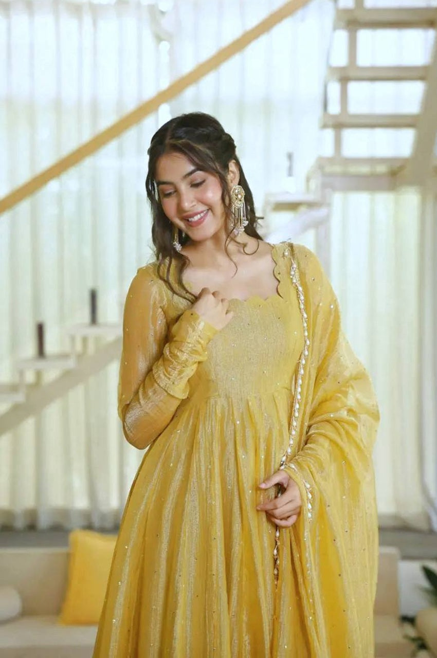 A woman wearing a bright yellow traditional outfit with a matching sheer dupatta, standing in a brightly lit indoor setting with a staircase in the background.
