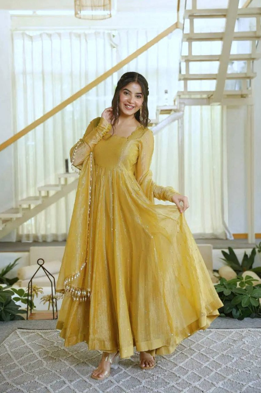 A woman modeling a flowing mustard yellow anarkali dress with sheer long sleeves and a matching embroidered dupatta, standing in a bright modern interior with white stairs and plants.
