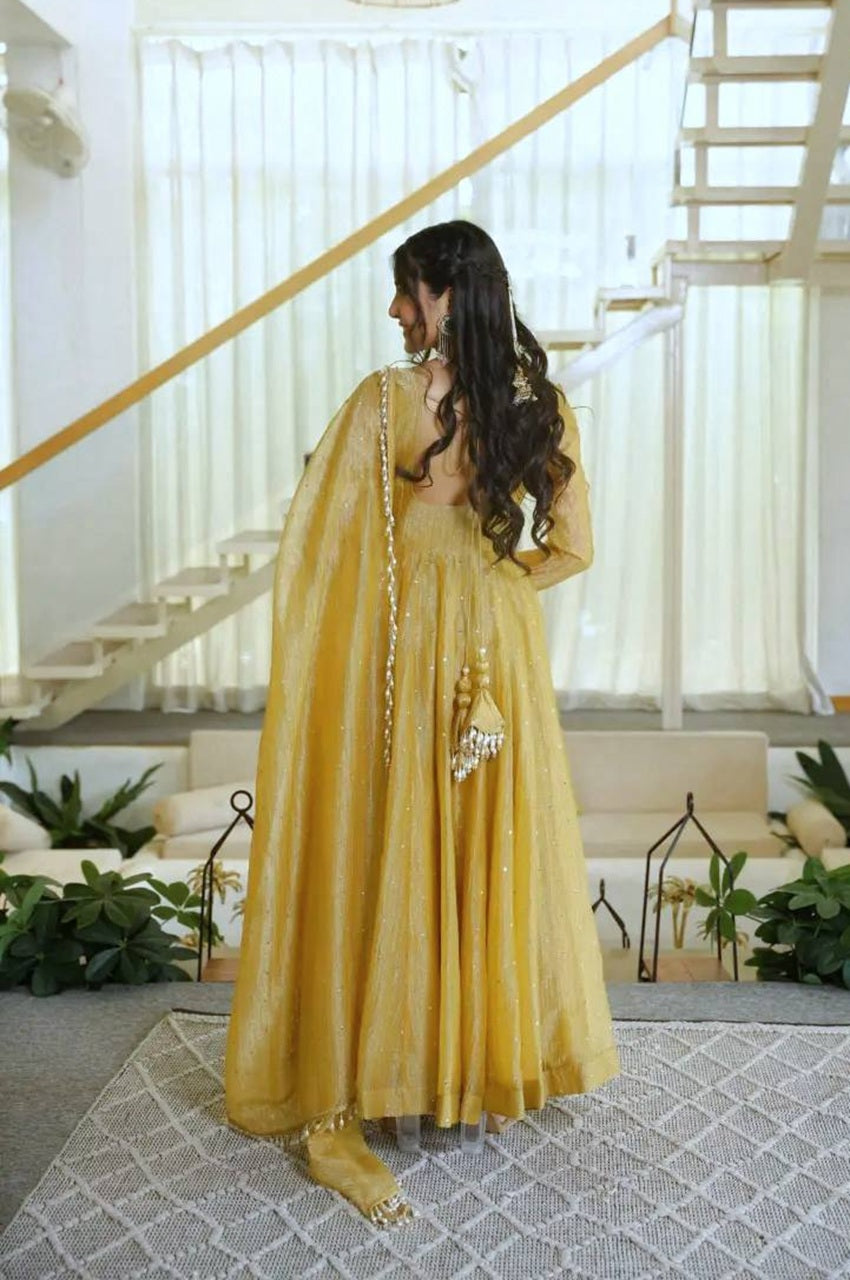 A woman wearing a flowing mustard yellow traditional dress with a sheer dupatta, standing in a bright indoor space with a staircase and plants in the background.