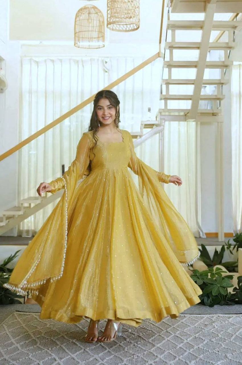 A woman wearing a flowing mustard yellow anarkali gown with sheer sleeves and a flared skirt, standing in a bright modern interior with a staircase and hanging lanterns.