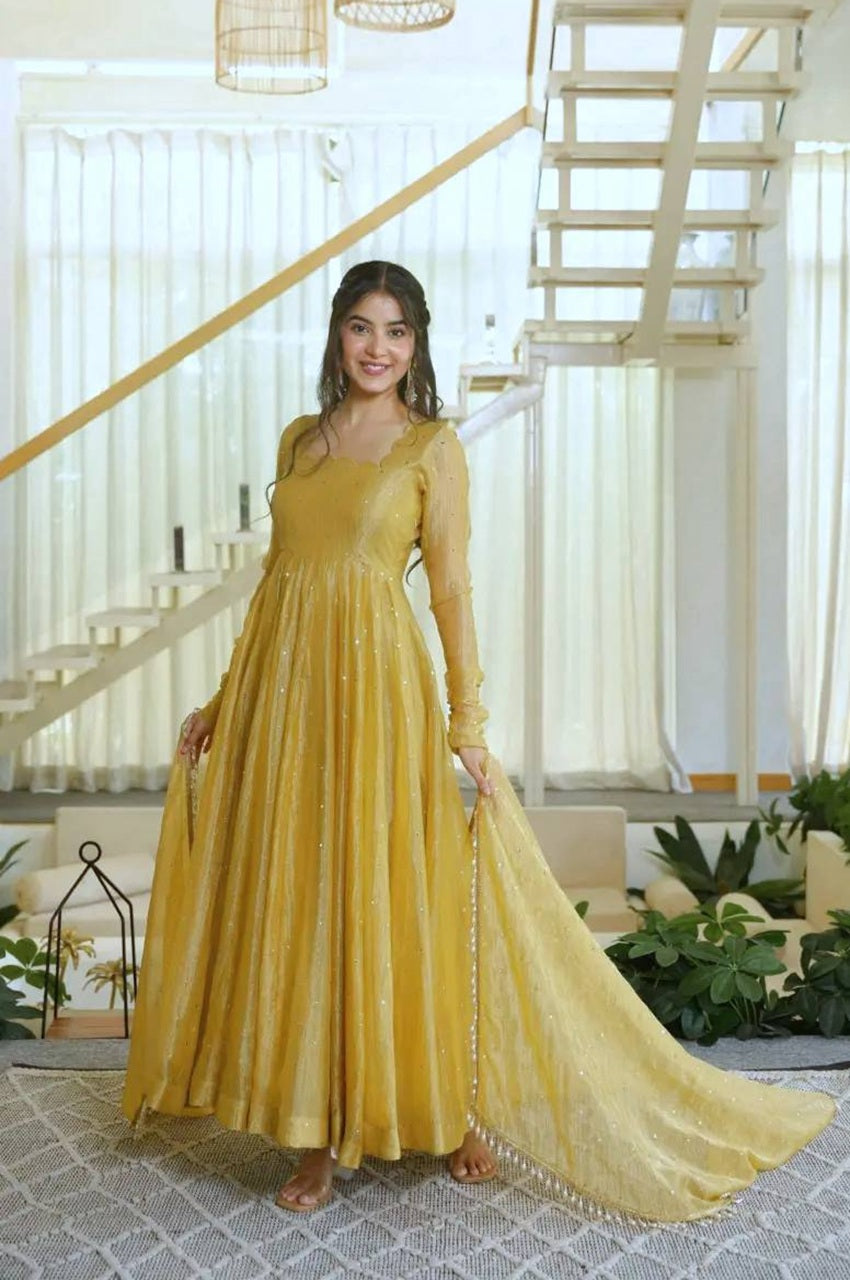 A woman wearing a long-sleeved mustard yellow anarkali gown with a matching dupatta, standing in a bright, modern room with a staircase in the background.