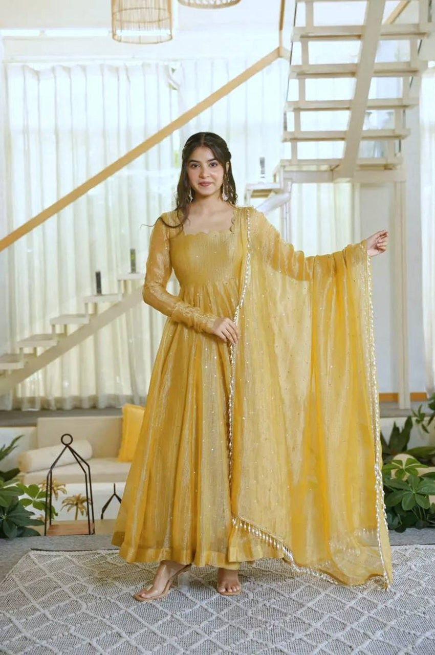 A woman models a mustard yellow traditional Indian anarkali gown with a matching sheer dupatta in a bright, modern indoor setting with a staircase in the background.