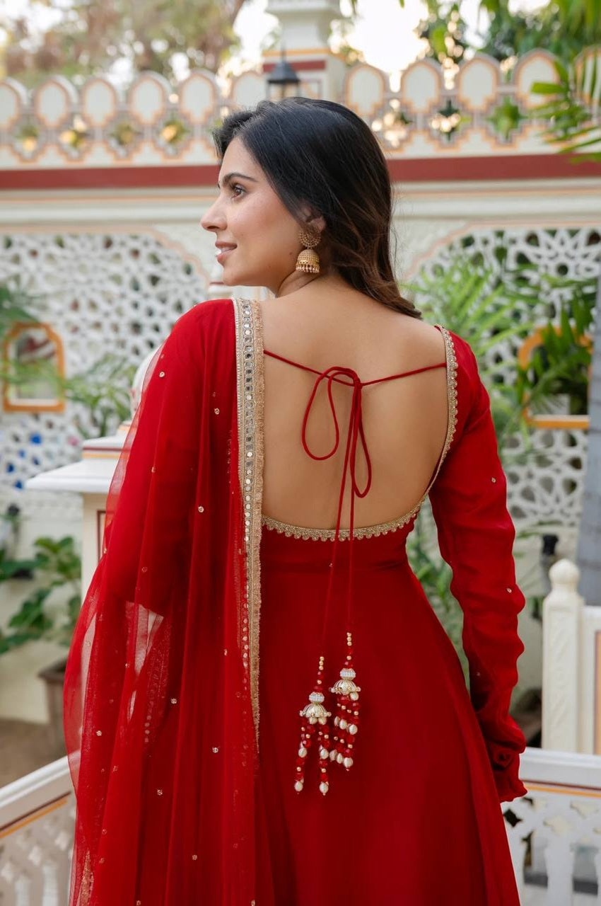 A woman wearing a red traditional dress with a deep open back, red tie straps, and a matching sheer dupatta with gold embroidery and tassel details, photographed outdoors in front of a decorative white railing and greenery.