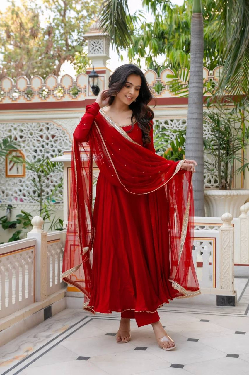 A woman modeling a traditional red Indian salwar kameez set with a matching dupatta, standing on an outdoor terrace with a decorative white railing and green plants in the background.