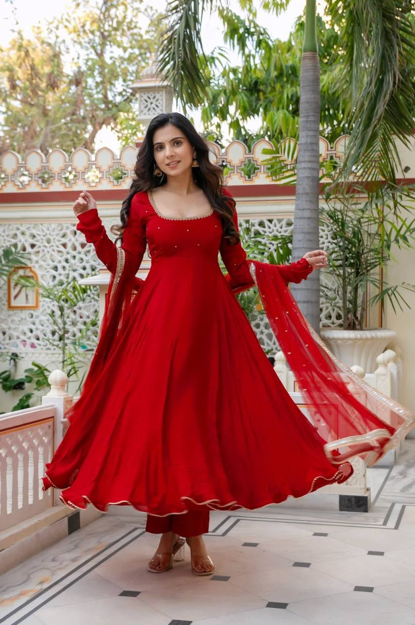 Woman in a red traditional outfit standing in an outdoor setting with greenery.