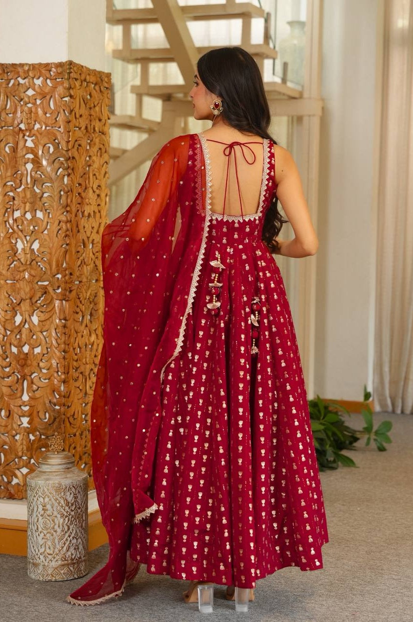 A woman wearing a deep red Anarkali gown with gold geometric patterns and a matching sheer dupatta, shown from the back in an indoor setting with a wooden staircase.