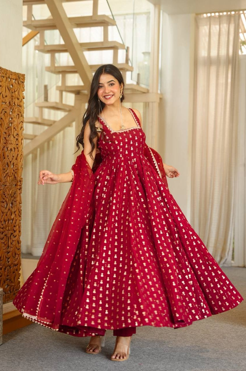 A woman modeling a red anarkali gown with white printed motifs in an indoor setting with a staircase and curtains in the background.