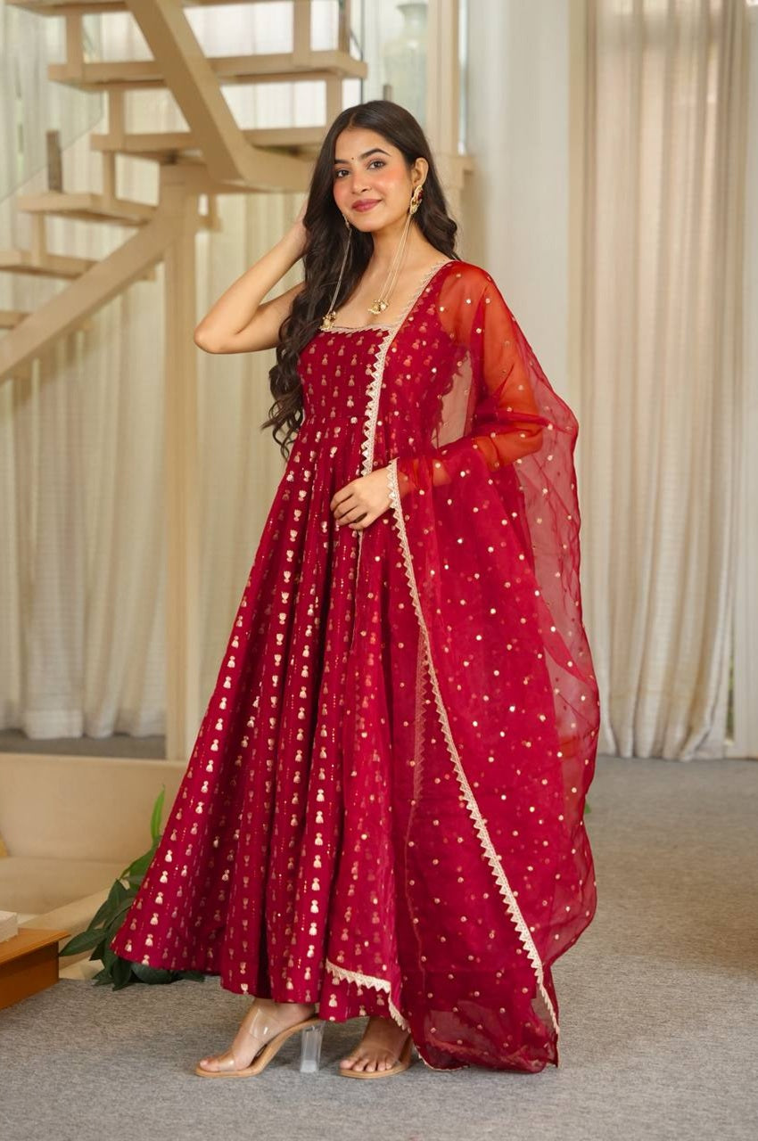 A woman models a deep red Anarkali gown with gold embroidery and a matching sheer dupatta in an indoor setting with a staircase and curtains.