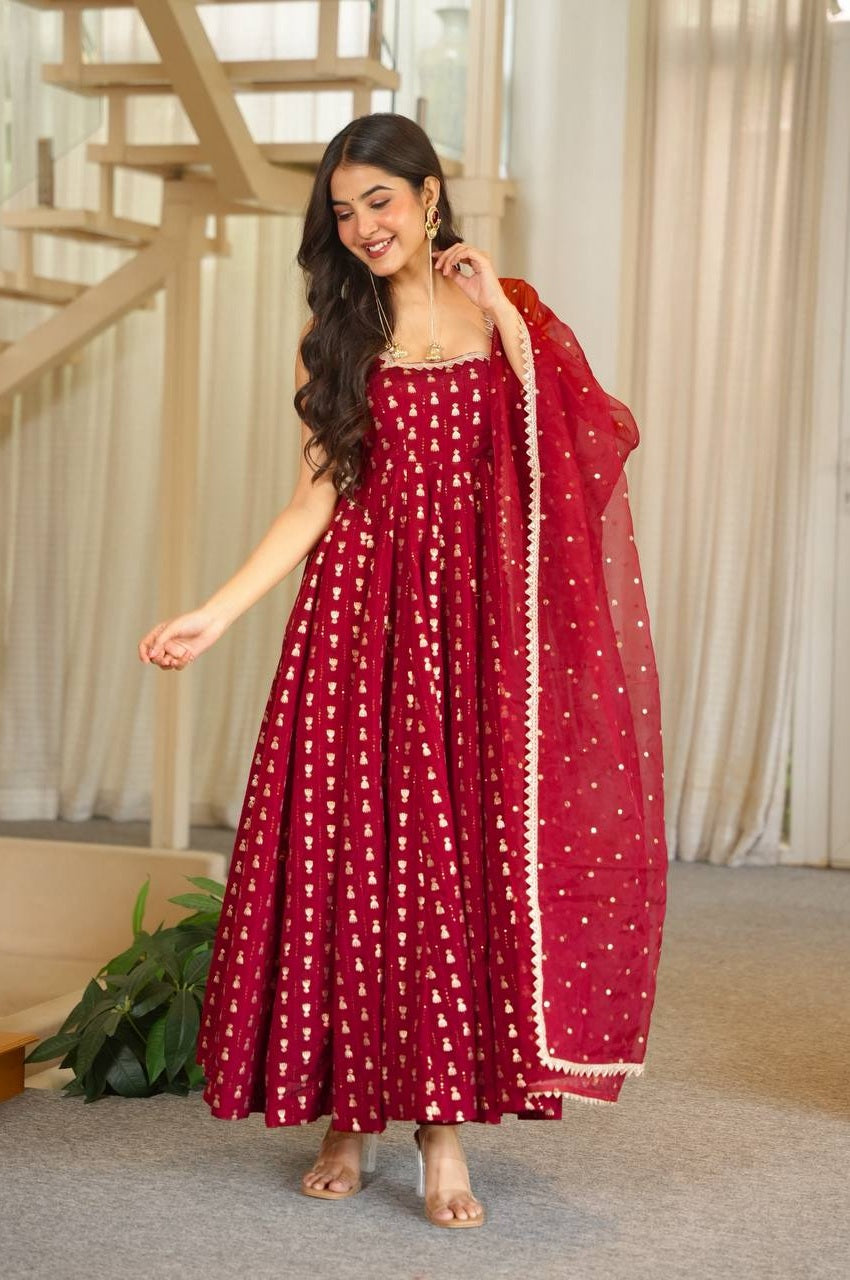A woman modeling a deep red anarkali gown with gold foil prints and a matching sheer dupatta draped over her shoulder, set in a bright indoor room with a staircase and curtains.