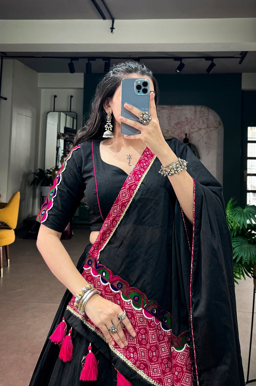 A woman takes a mirror selfie wearing a black saree with a vibrant magenta and green border pattern and pink tassels. She is wearing silver jewelry, including a necklace, earrings, and bracelets, and holding a blue smartphone.