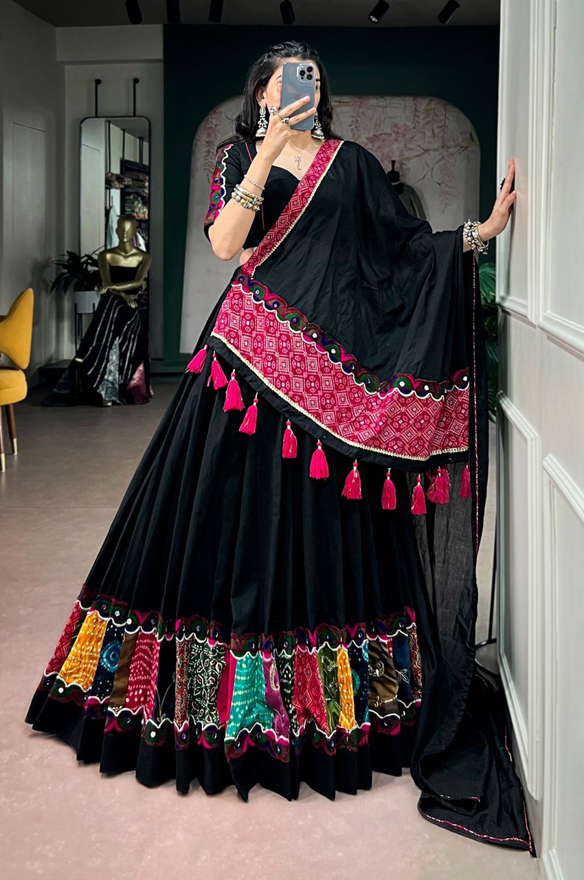 A woman takes a mirror selfie wearing a black traditional dress with a wide pink and multicolor embroidered border and tassel details. The setting appears to be an indoor boutique or fitting space with a mirror, wall decor, and clothing visible in the background.