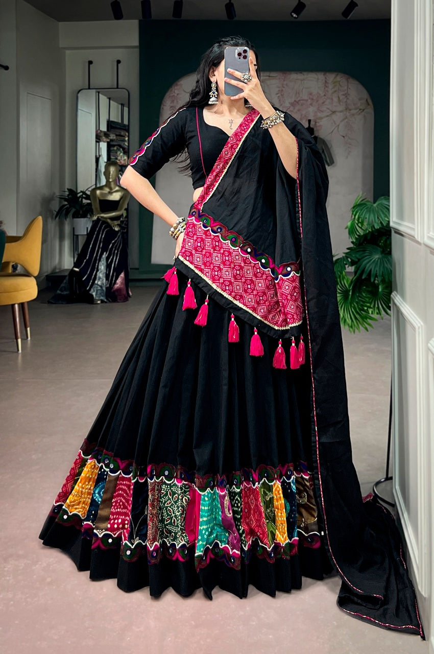 A woman takes a mirror selfie wearing a black traditional Indian lehenga choli with vibrant pink, red, green, and gold embroidered borders and tassels, set in an indoor boutique environment.