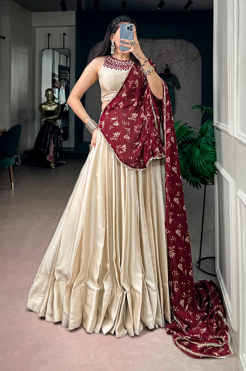A woman takes a mirror selfie wearing a cream-colored satin lehenga choli with a deep maroon dupatta featuring gold floral patterns. The setting appears to be a boutique or dressing room with a mannequin and plants in the background.