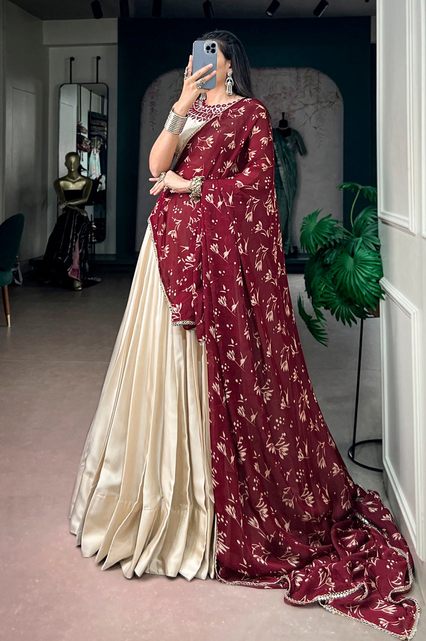 A woman takes a mirror selfie in a boutique wearing a traditional Indian saree. The saree features a deep maroon-red border with a gold floral pattern and a flowing cream-colored body.