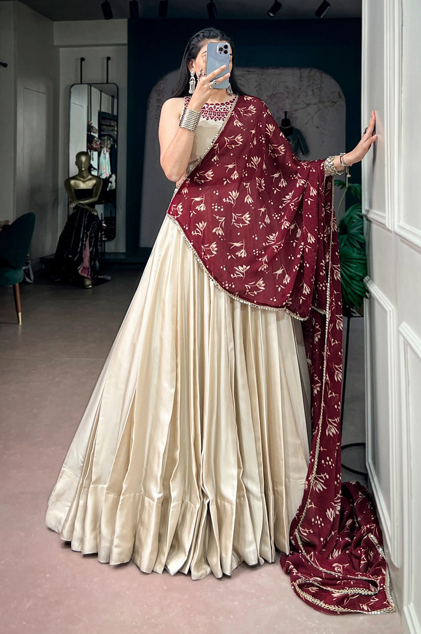 A woman takes a mirror selfie wearing a traditional Indian outfit consisting of a cream-colored pleated skirt (lehenga) and a maroon dupatta with gold floral patterns and embroidery.
