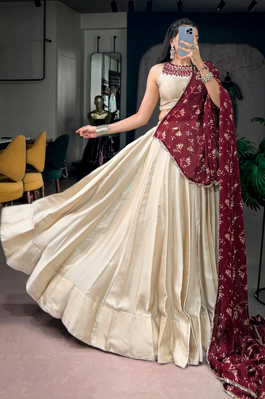 A woman takes a mirror selfie in an indoor boutique setting, wearing a cream-colored sleeveless lehenga choli with a deep neckline and a flowing skirt, paired with a maroon dupatta featuring gold floral patterns.