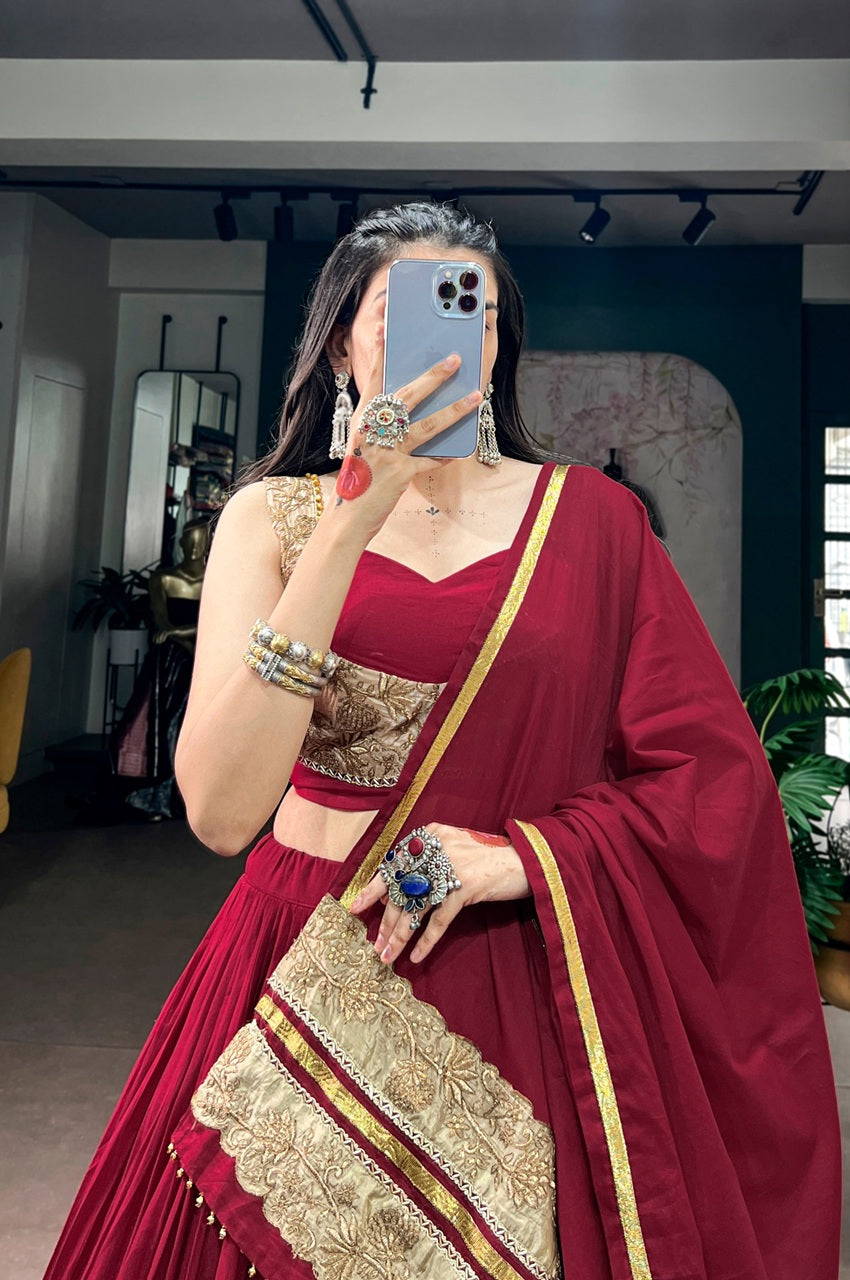 A woman takes a mirror selfie wearing a deep red Indian ethnic outfit with gold embroidery and a matching dupatta. The setting appears to be a mirror selfie in an indoor room with home decor elements visible in the background.