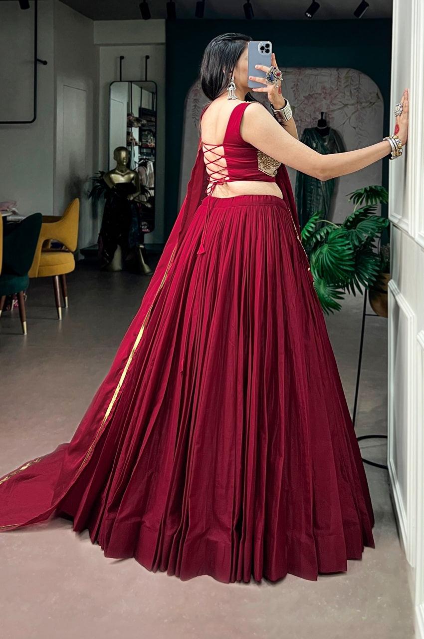 A woman takes a mirror selfie in an indoor boutique setting, wearing a deep maroon red traditional Indian lehenga choli dress with a pleated skirt, lace-up back, and a matching dupatta with gold border details.