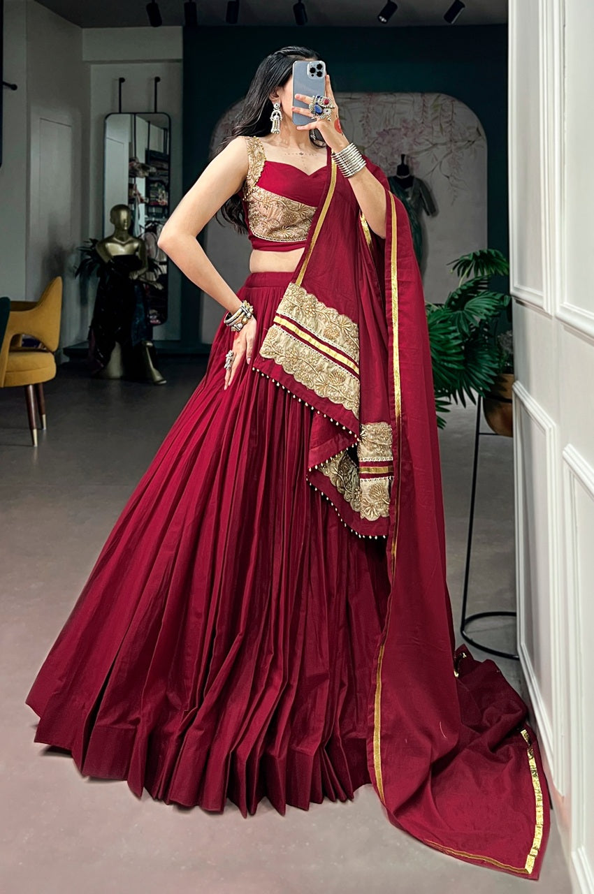 A woman takes a mirror selfie wearing a deep maroon lehenga choli with gold embroidery and a matching dupatta with gold borders, set in an indoor boutique environment.