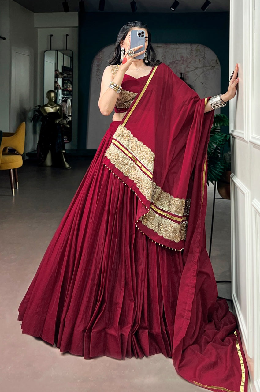 A woman takes a mirror selfie wearing a voluminous deep maroon red lehenga choli with gold embroidered borders and a matching dupatta, set in an indoor boutique environment.