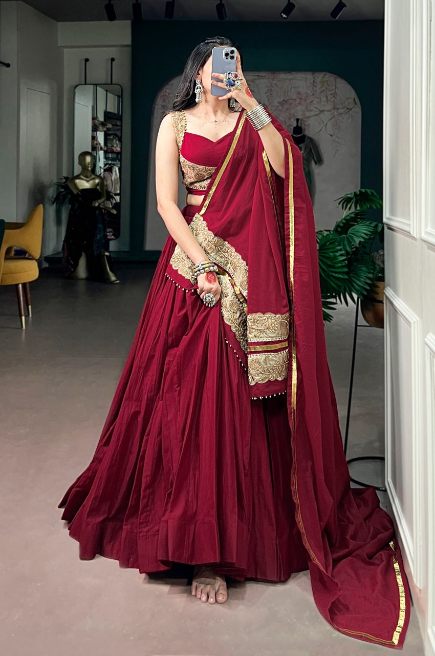 A woman takes a mirror selfie wearing a traditional Indian lehenga choli outfit in deep maroon with gold embroidery and a matching dupatta with a gold border, set in an indoor boutique environment.