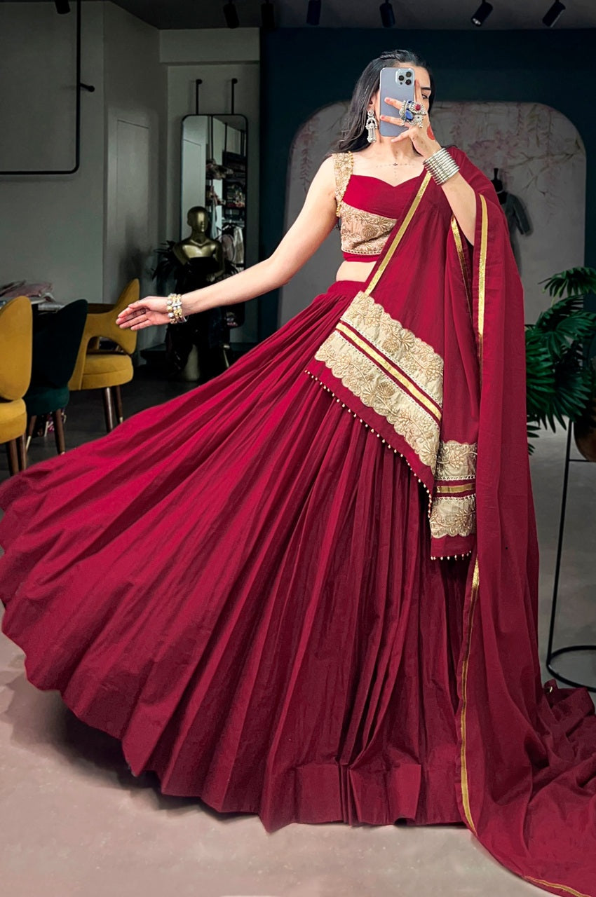 A woman wearing a deep maroon red lehenga choli with gold embroidery, taking a mirror selfie in an indoor studio setting.