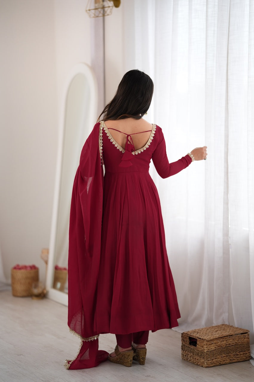 A woman viewed from behind wearing a long-sleeved deep red traditional dress with a draped dupatta and decorative gold trim on the neckline, standing in a bright room with white curtains.