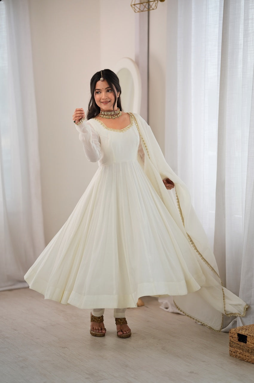 A woman modeling a long white Anarkali gown with a matching sheer dupatta draped over her shoulders, standing in a bright room with white curtains.