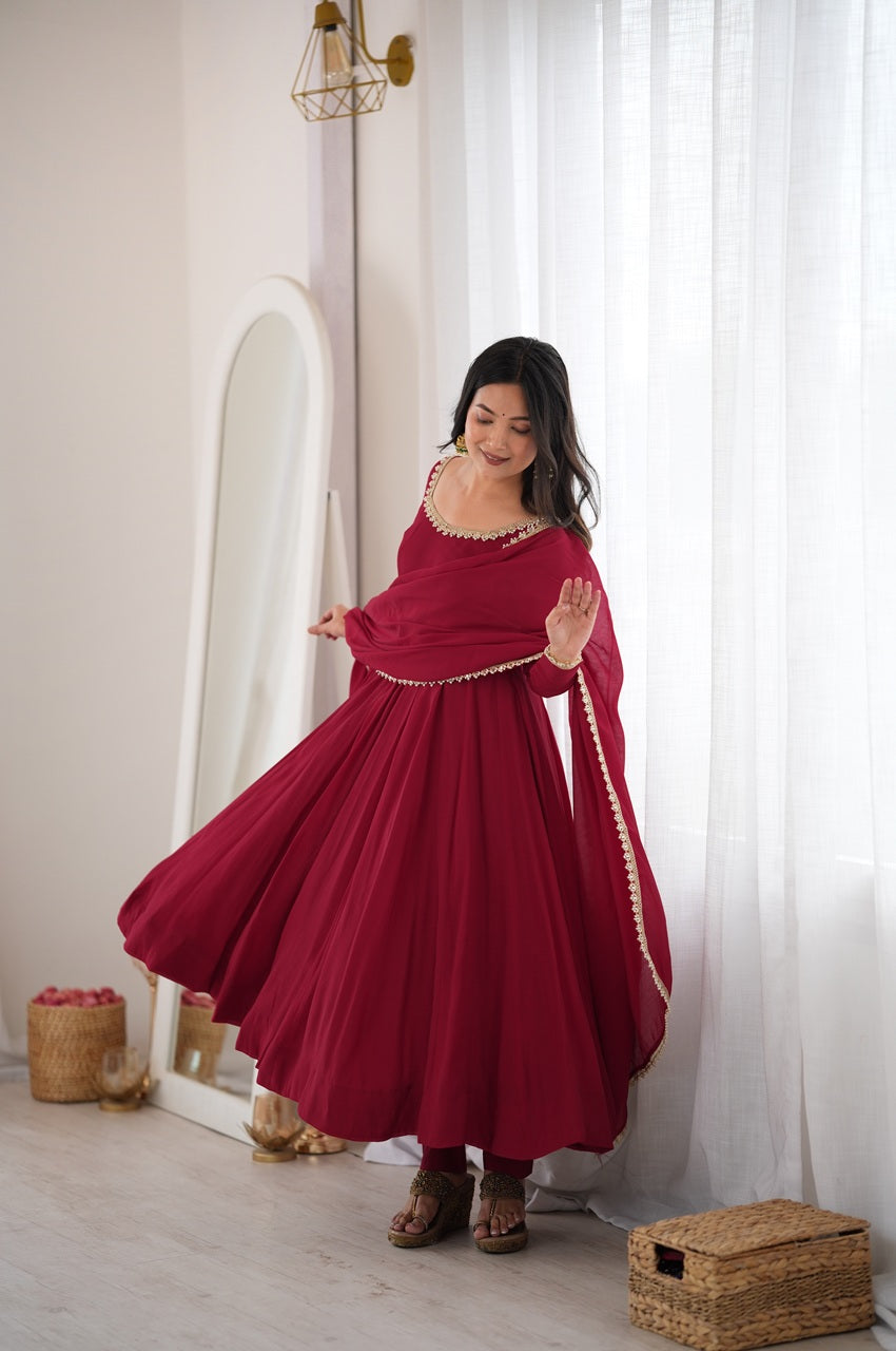 A woman wearing a deep red Anarkali-style dress with gold embroidery and a matching dupatta, standing in a bright room with sheer white curtains, a mirror, and woven baskets.