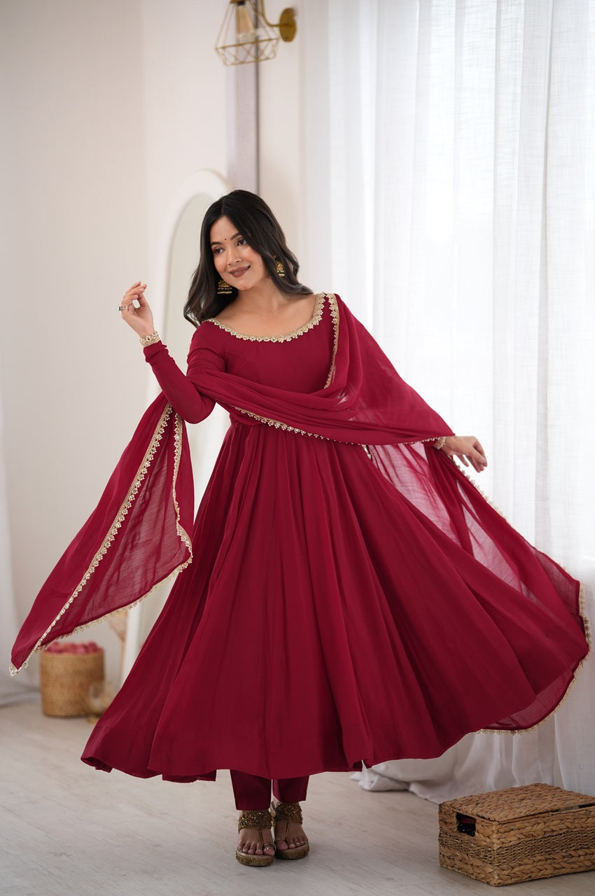 A woman modeling a deep red long-sleeve Anarkali gown with a matching dupatta and red trousers, standing in a bright, white-walled room with sheer curtains.