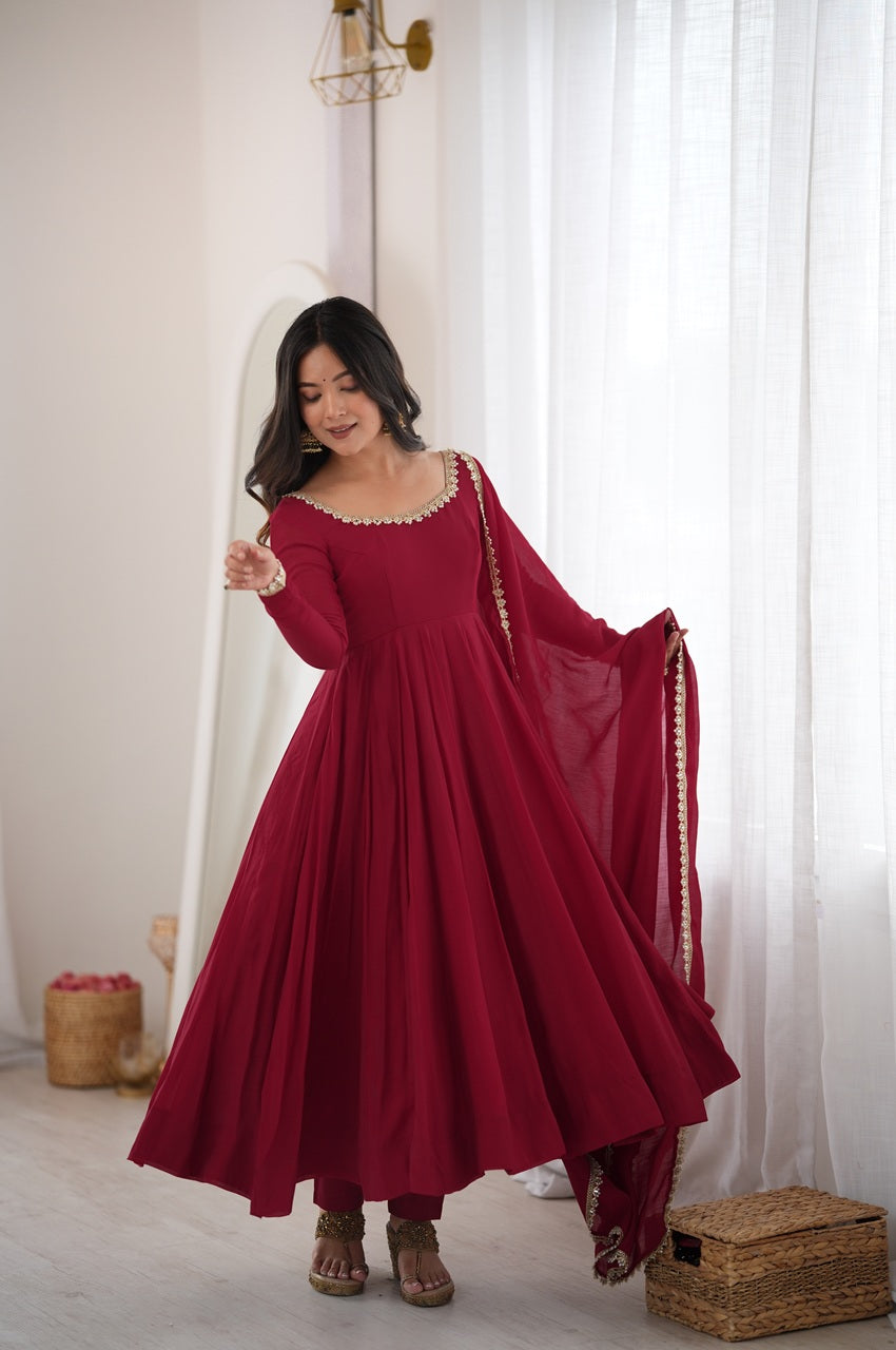 A woman wearing a long-sleeved deep red Anarkali gown with gold embroidery and a matching dupatta, standing in a bright room with white curtains.