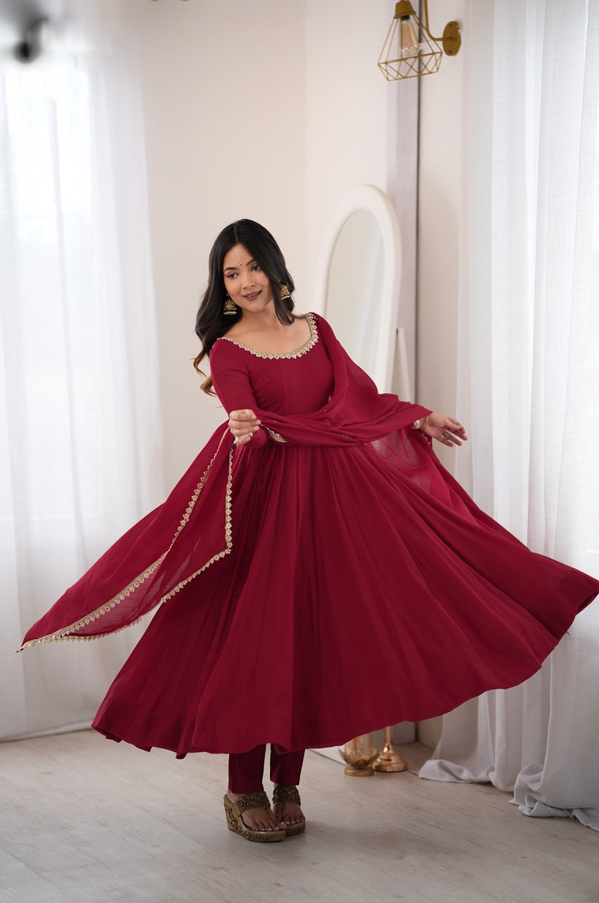 A woman wearing a flowing deep red anarkali-style dress with gold-trimmed neckline and dupatta, standing in a bright room with white curtains and a mirror.